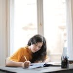 A young woman focused on writing in her notebook near a sunlit window.