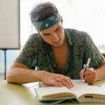 Young man intensely studying with a notebook at a desk indoors, focusing on research and writing.